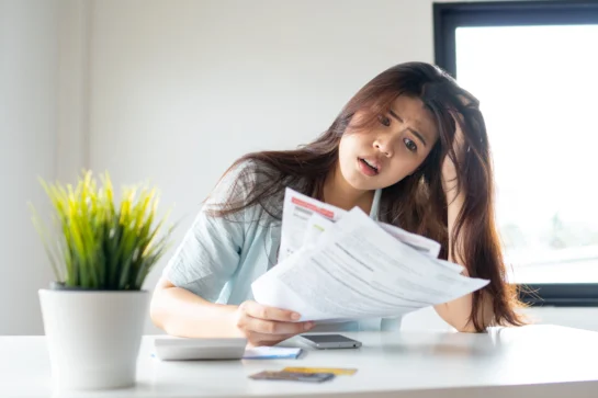 Como gerar 2ª via de boleto vencido em poucos minutos? 1 Mulher preocupada analisando papéis e boletos, buscando a 2ª via de boleto em uma mesa com um vaso de planta e celular.