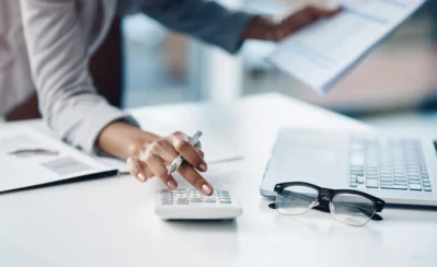 Pessoa usando uma calculadora, com laptop e óculos ao lado. A pessoa está lendo sobre o que é split de pagamento e como facilitar esse processo.