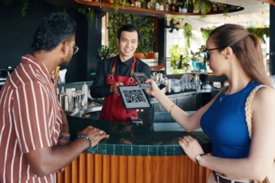 Casal em restaurante no caixa realizando pagamento para o garçom. A mulher mirando celular em QR code dinâmico e estático para pagar.