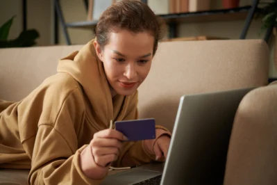 Mulher deitada no sofá, segurando um cartão e preenchendo informações no computador. Ela está aprendendo a como controlar vendas no cartão de crédito.