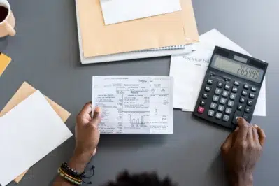 Mãos segurando uma conta enquanto usa uma calculadora. Na mesa, há envelopes e papéis, mostrando a importância de saber como vender no boleto parcelado.