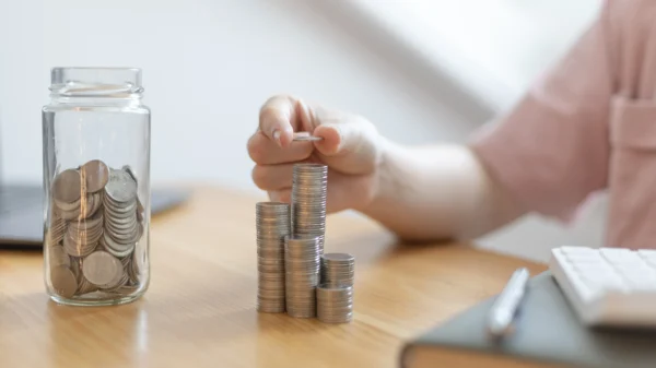 10 dicas de como economizar dinheiro e cortar gastos 1 Mulher está aprendendo como economizar dinheiro, empilhando moedas em cima de uma mesa e guardando em um pote.