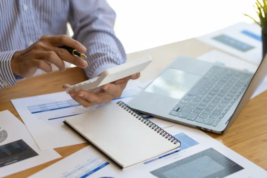 Aprenda como planejar a aposentadoria com essas 6 dicas essenciais 1 Homem fazendo contas de como planejar a aposentadoria na calculadora. Papéis e computador estão sobre a mesa.