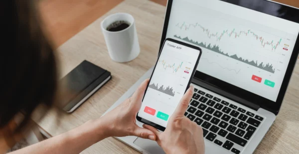 O que é investimento financeiro? Tudo o que você precisa saber 1 Pessoa usando um aplicativo de investimentos no celular e no notebook, ilustrando o que é investimento financeiro.
