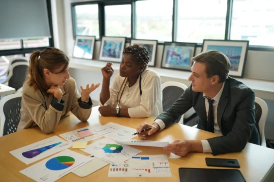 Reunião com três pessoas discutindo gráficos e relatórios financeiro. Eles estão estudando como fazer um planejamento financeiro empresarial.